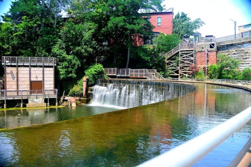 A serene waterway with a gentle waterfall, surrounded by greenery and historic buildings under a clear blue sky.