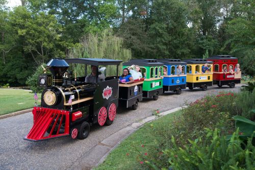 A colorful miniature train with passengers rides along a scenic path surrounded by greenery.