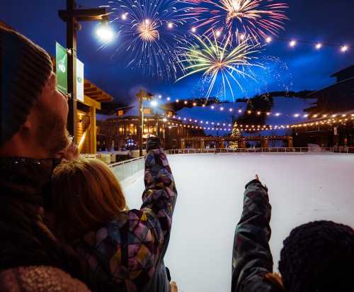 A family watches fireworks over an ice skating rink, pointing at the colorful display against a night sky.