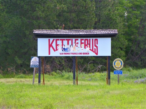 Sign for Kettle Falls, featuring the slogan "1640 Friendly People & One Grouch," surrounded by greenery.
