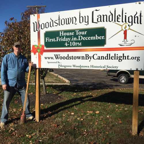 A man stands next to a sign for "Woodstown by Candlelight," promoting a December house tour.