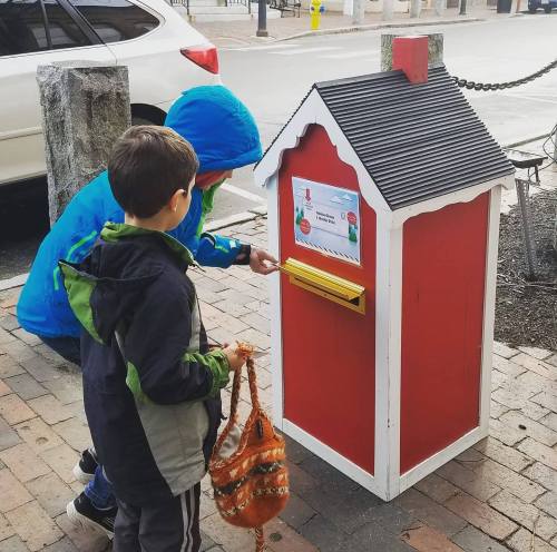 Two children in colorful jackets are approaching a red mailbox with a black roof, preparing to drop something inside.