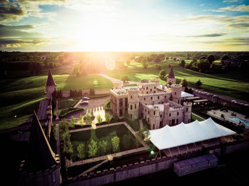 Aerial view of a castle with towers, surrounded by green fields and a large tent, under a bright sunset sky.