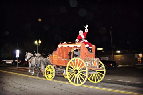 Santa Claus waves from a festive horse-drawn carriage, illuminated by streetlights on a dark night.