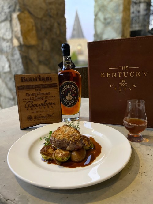 A gourmet dish with roasted vegetables, a bourbon bottle, and a glass of bourbon, set against a castle backdrop.