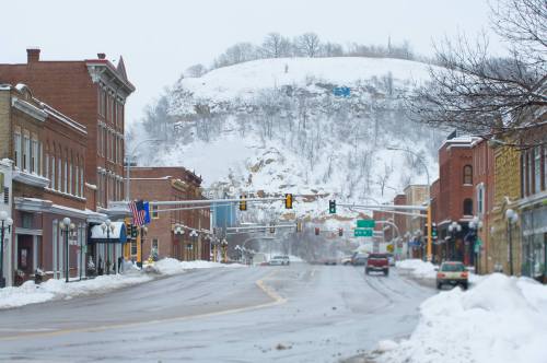 Snow-covered street in a small town, with historic buildings and a hill in the background. Traffic lights visible.