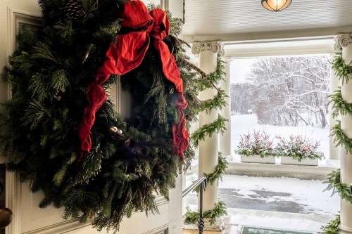 A festive wreath with a red bow hangs on a door, surrounded by snowy scenery and decorated greenery.