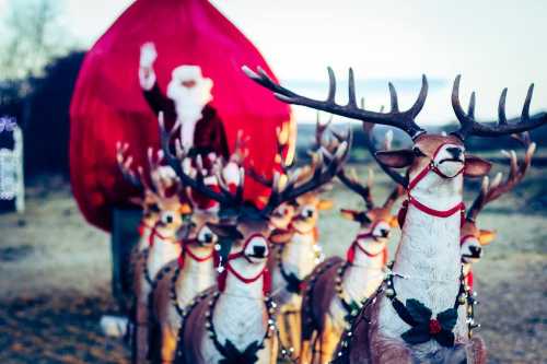 A line of reindeer with festive decorations, with Santa waving in the background near a red tent.