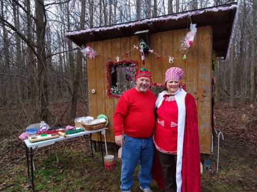 A couple in festive attire stands in front of a decorated wooden cabin in a forest, surrounded by holiday decorations.