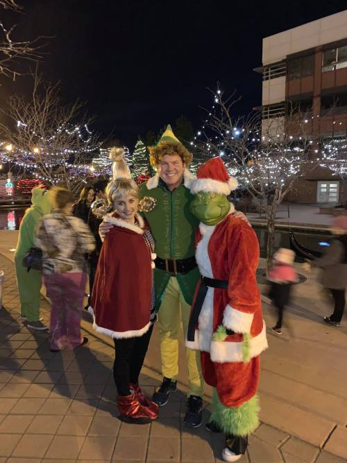 A festive scene with three people dressed as the Grinch, an elf, and a holiday character, surrounded by holiday lights.