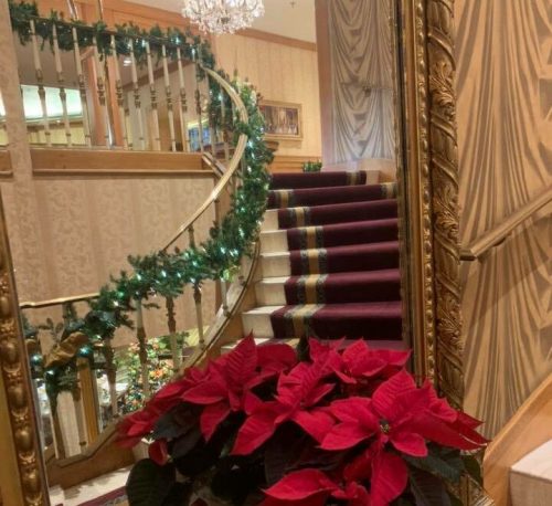 A festive scene featuring red poinsettias in a pot, a mirrored wall, and a staircase adorned with garland and a chandelier.