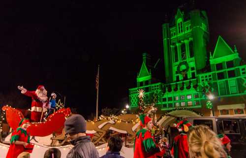 A festive parade scene with Santa on a sleigh, surrounded by elves and a brightly lit building in the background.