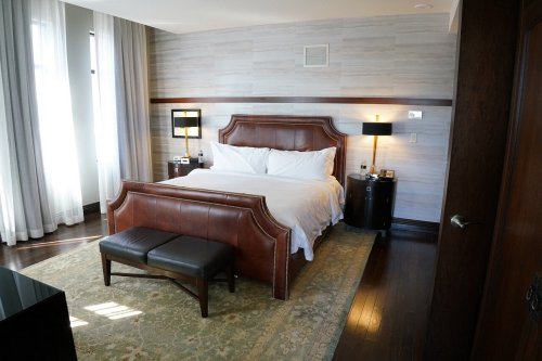 A stylish hotel room featuring a large bed, bedside tables, and a bench, with large windows and elegant decor.