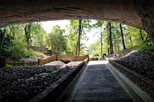 A pathway leads through a rocky area under a stone arch, surrounded by trees and natural light.