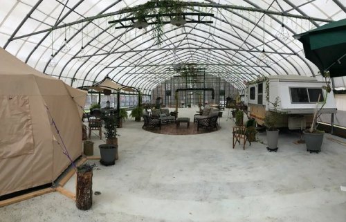 A spacious greenhouse interior with seating areas, plants, and a tent on one side. Natural light filters through the roof.