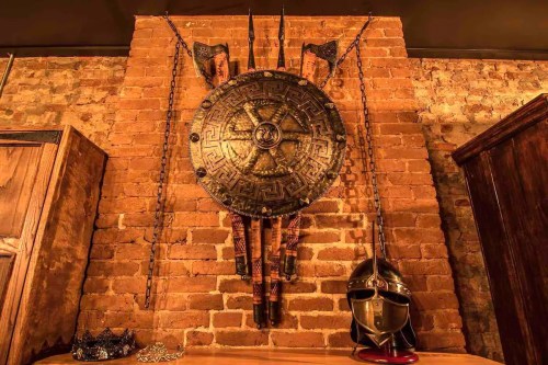 A decorative wall display featuring a large shield, spears, and a helmet against a brick background.