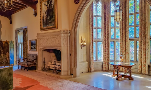 A spacious room with a stone fireplace, large windows, and elegant decor, featuring a wooden table and artwork on the walls.