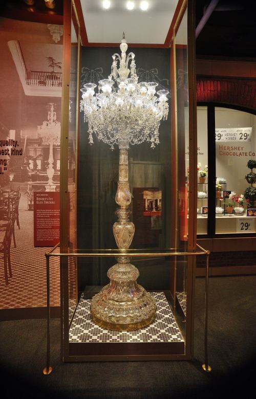 A large, ornate crystal chandelier displayed in a glass case, showcasing intricate details and sparkling lights.