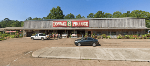 A rustic produce store with a large sign reading "Donna's 6 Produce," surrounded by greenery and parked cars.