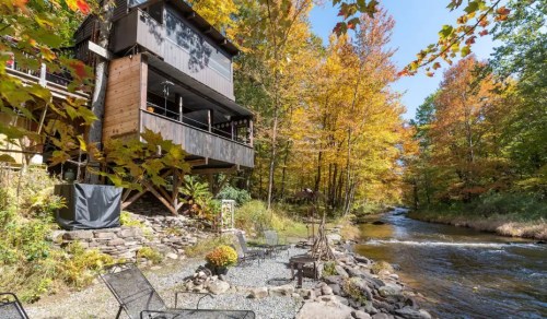 A modern cabin by a river, surrounded by vibrant autumn foliage and outdoor seating by the water.