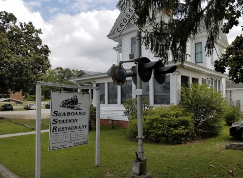 Sign for Seaboard Station Restaurant next to a railroad crossing, with a white house and greenery in the background.
