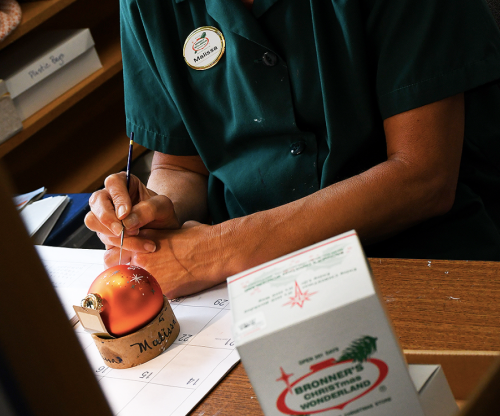 A person in a green shirt decorates an orange ornament at a wooden table, with a box labeled "Bronner's Christmas Wonderland."