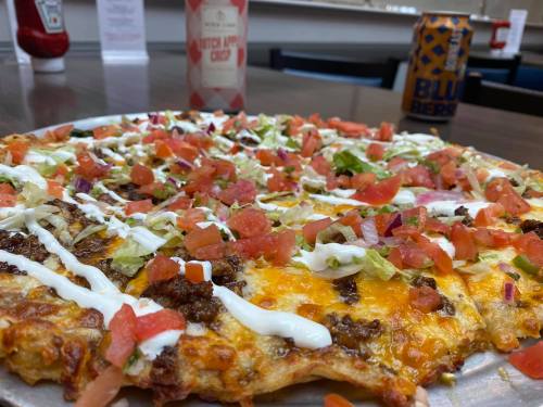 A close-up of a pizza topped with ground beef, tomatoes, lettuce, onions, and drizzled with sour cream, alongside drinks.