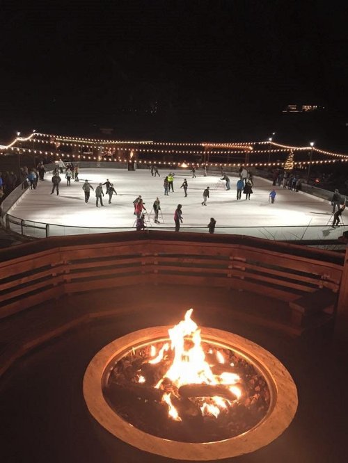 A nighttime ice skating rink filled with skaters, illuminated by string lights, with a fire pit in the foreground.