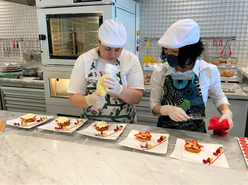 Two chefs in white hats and gloves decorate desserts on a marble countertop, focusing on plating with fruits and sauces.