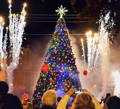 A brightly lit Christmas tree adorned with colorful ornaments, surrounded by fireworks and a crowd celebrating.