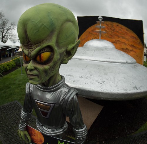 A green alien figure stands next to a silver UFO, with a large planet backdrop and cloudy sky.