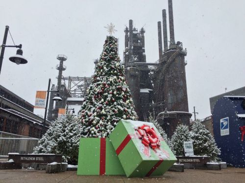 A decorated Christmas tree with large gift boxes in front of an industrial backdrop, lightly snowing.