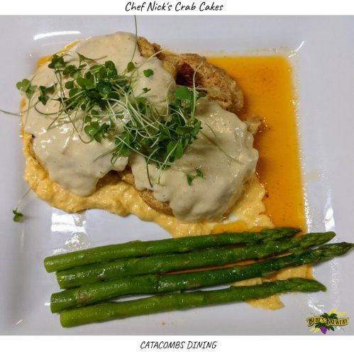 Plate of crab cakes topped with creamy sauce, served on a bed of grits, with asparagus on the side.