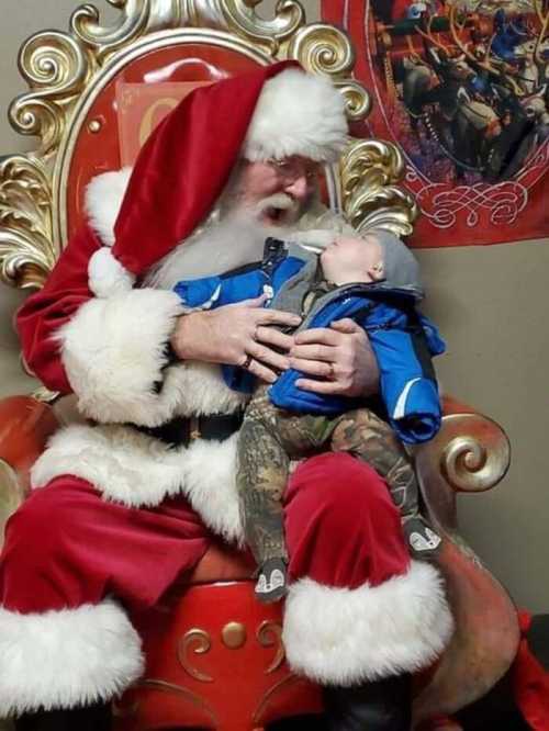 Santa Claus in a red suit holds a baby in a blue jacket, both looking at each other, seated on a festive throne.