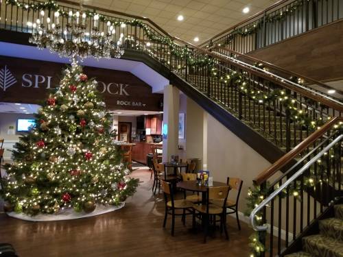 A festive lobby with a decorated Christmas tree, stairs adorned with lights, and dining tables nearby.