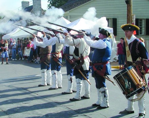 Historical reenactors in period costumes fire muskets during a demonstration, with a crowd watching in the background.