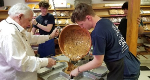An older man and a young boy pour a large mixture into baking trays in a bakery.