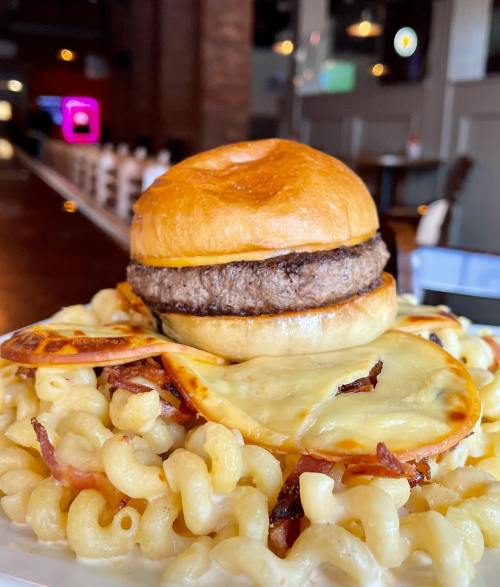 A close-up of a burger on macaroni and cheese, topped with crispy bacon and melted cheese, served in a restaurant setting.