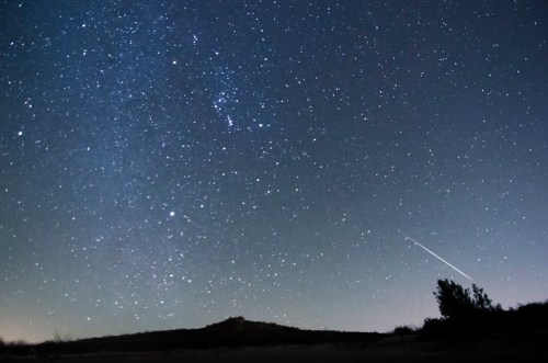 A starry night sky filled with stars, featuring a shooting star and a silhouette of a distant hill.