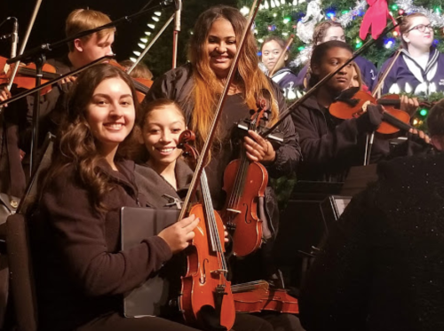 A group of musicians smiling while holding violins, performing in front of a festive backdrop with a Christmas tree.