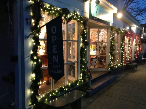 A festive gallery storefront decorated with lights and greenery, featuring an "OPEN" sign in the evening.