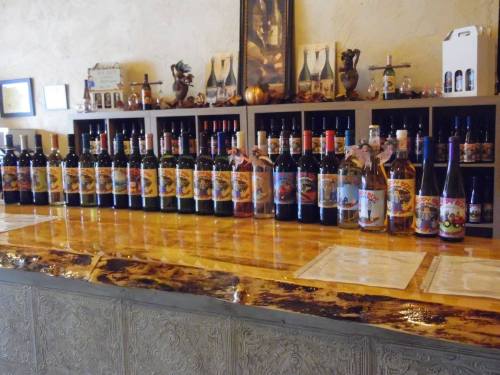 A display of various wine bottles lined up on a wooden counter, with shelves of more bottles in the background.