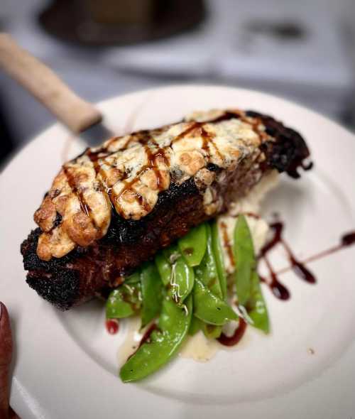 A plate featuring a grilled steak topped with cheese, served with sautéed snap peas and a drizzle of sauce.