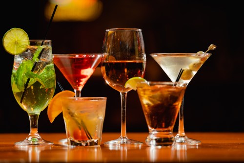 A variety of colorful cocktails in elegant glasses, featuring garnishes like lime and orange slices, against a dark background.