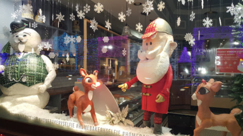 Holiday window display featuring a snowman, Santa, and reindeer with snowflakes in the background.