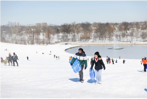 Two people walk up a snowy hill with sleds, while others enjoy winter activities around a frozen pond.