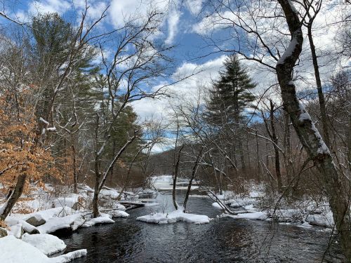 A serene winter landscape featuring a snow-covered river, bare trees, and a cloudy blue sky.