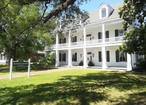 A large white two-story building with a porch, surrounded by green grass and trees, featuring a sign in front.