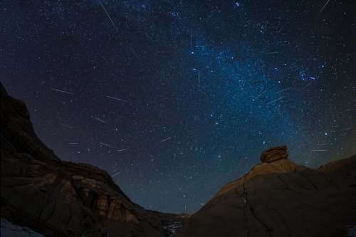 A starry night sky filled with shooting stars over rugged rock formations.