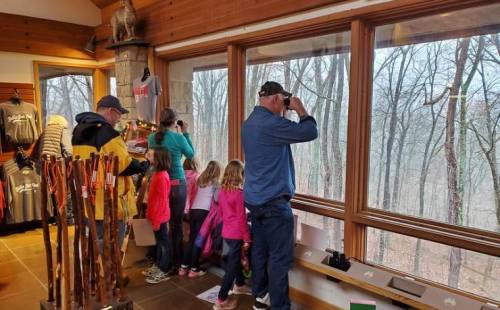 A group of children and adults observe nature through large windows in a cozy indoor space.